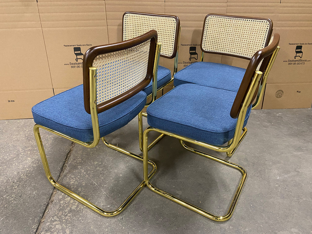 Four mid-century modern dining chairs with blue upholstered seats and backs, woven cane back panels, and curved gold metal frames, arranged on a concrete floor in front of cardboard boxes.