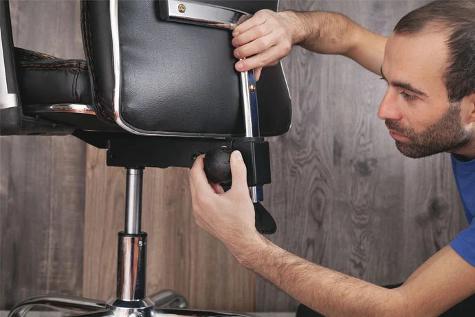 A man adjusting the gas lift and castor wheel of a black leather office chair with chrome accents, set against a dark wood-paneled wall.